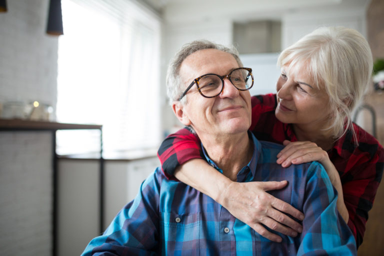 Smiling older woman hugging an older man in glasses from behind at home