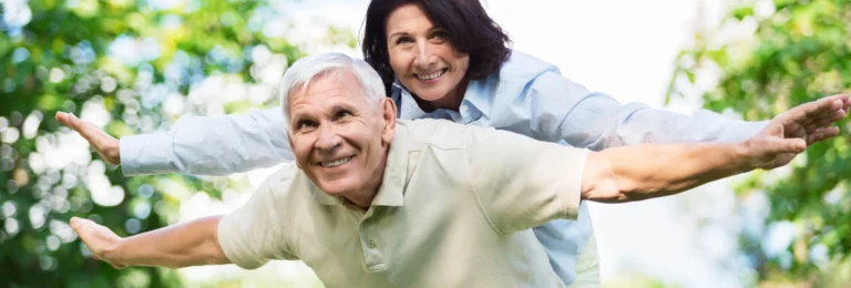 Smiling senior man with arms outstretched while a woman stands behind him outdoors